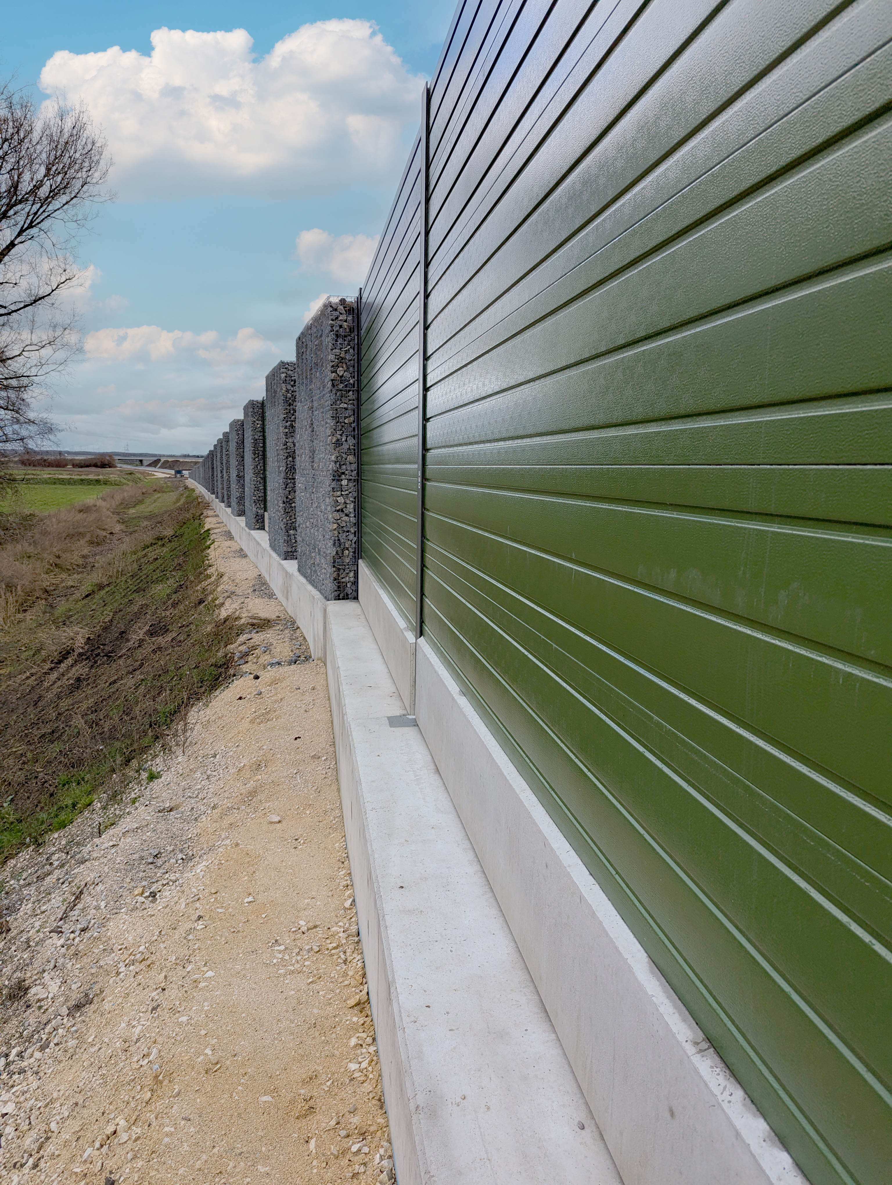 Gesamtansicht neu errichteter Lärmschutzwände in den Donauauen entlang der Querspange Erbach, mit farbigen Aluminiumelementen und Gabionen